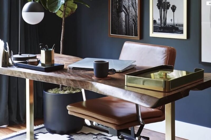 Tamni radni prostor sa masivnim drvenim stolom, kožnom stolicom, laptopom, šoljom kafe, radnom lampom, biljkama i uokvirenim fotografijama na zidu
