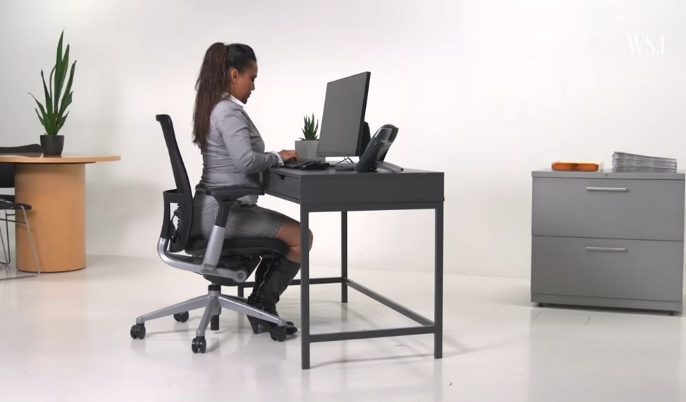 Osoba sedi za radnim stolom na ergonomskoj stolici i radi za racunarom u jednostavnom, svetlom prostoru