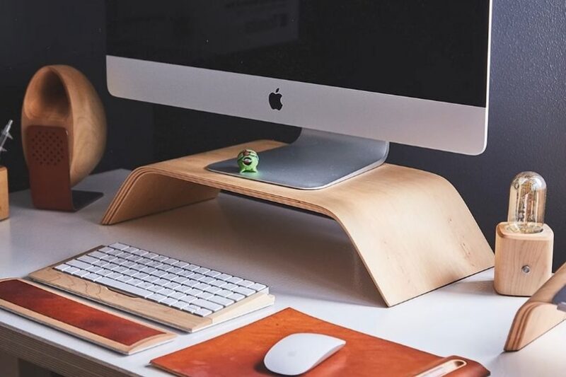 Minimalistički radni sto sa iMac računarom na drvenom postolju, tastaturom i mišem, kožnim podmetačem i malim dekorativnim predmetima