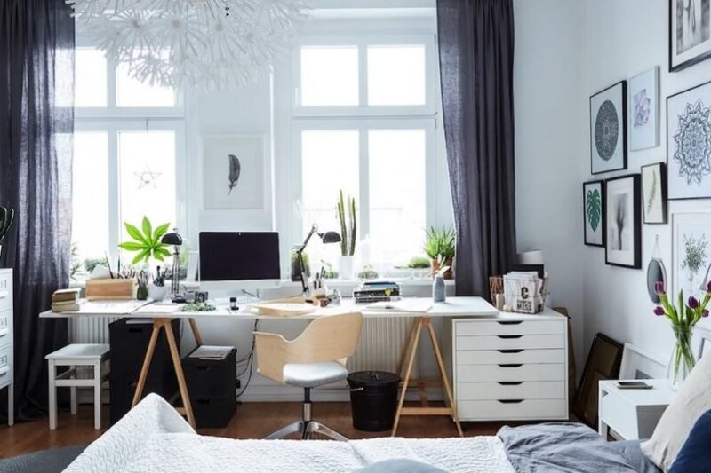 Svetla kućna kancelarija sa belim stolom ispred velikih prozora, radnim lampama, računarom, biljkama i galerijom uokvirenih slika na zidu